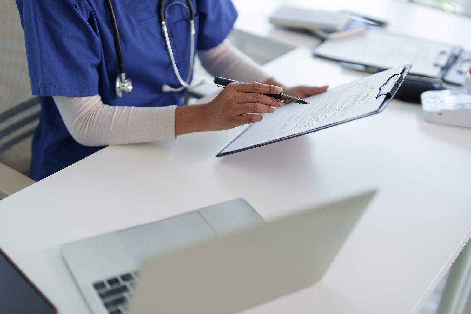 Manos de un profesional de la salud con ambo azul y estetoscopio señalando con una lapicera un formulario en un portapapeles, junto a una laptop