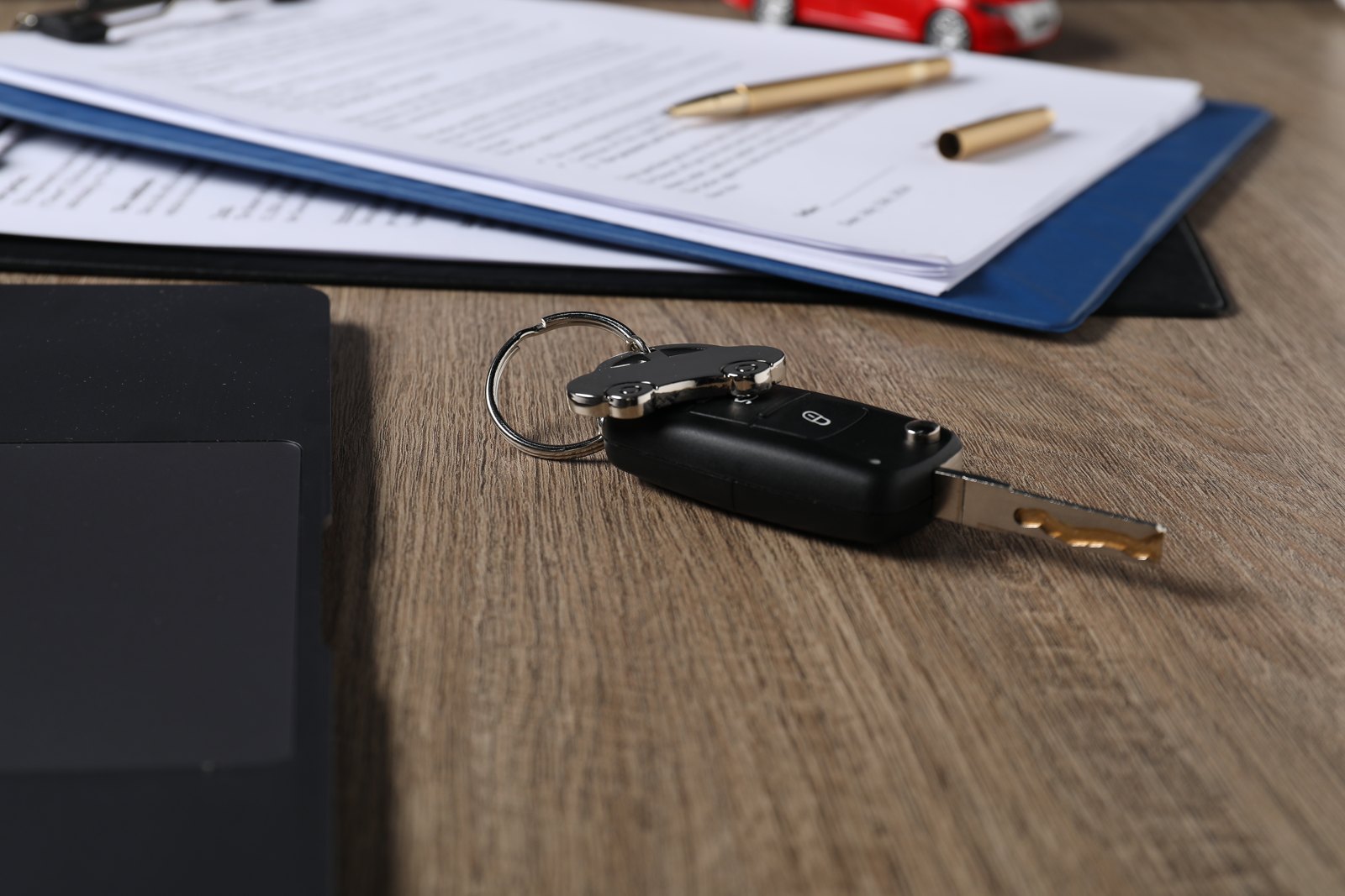 Llave de auto con control remoto sobre una mesa de madera, junto a una carpeta azul con documentos y una lapicera dorada